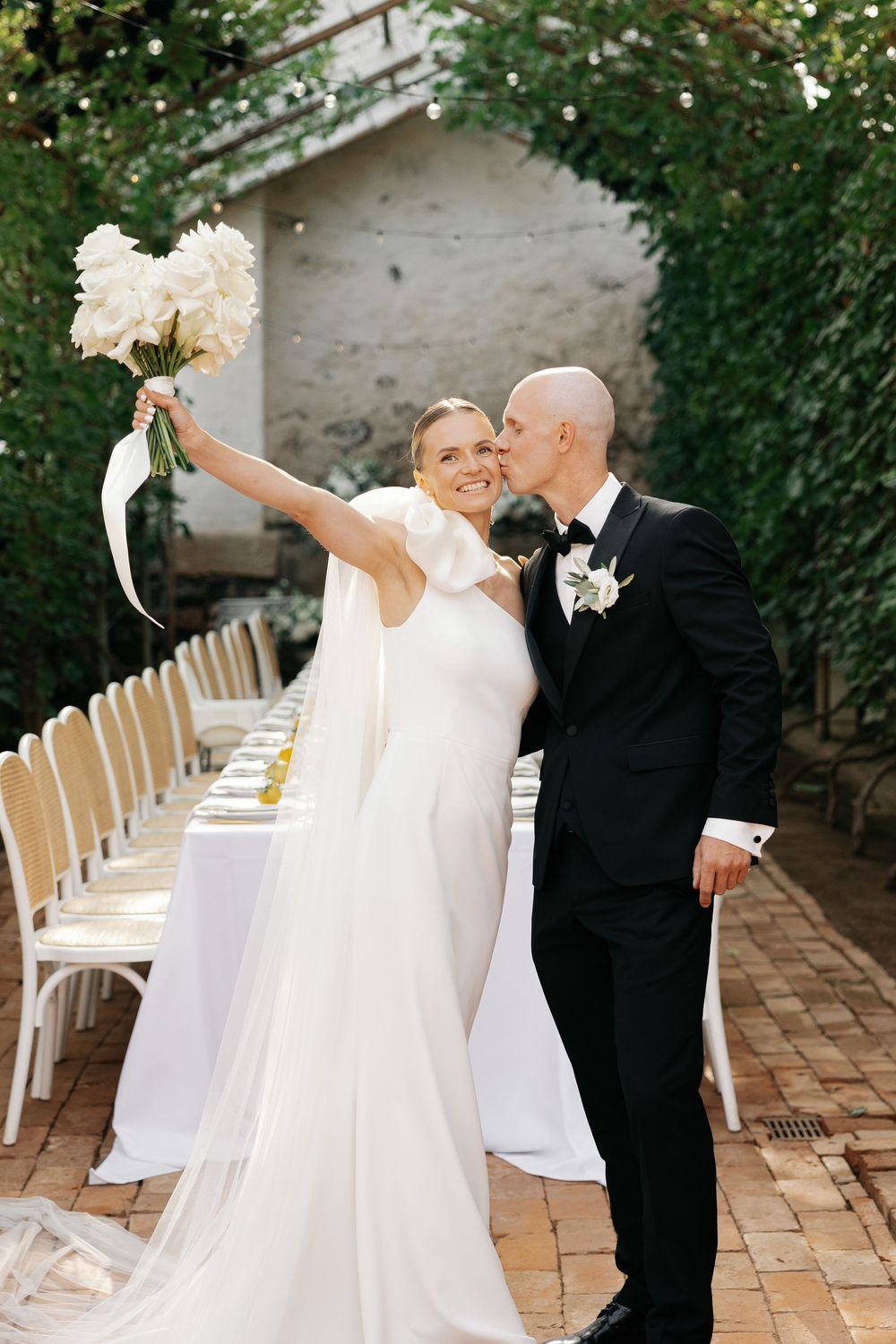 Newlyweds share romantic moment on brick patio with white chairs and string lights.
