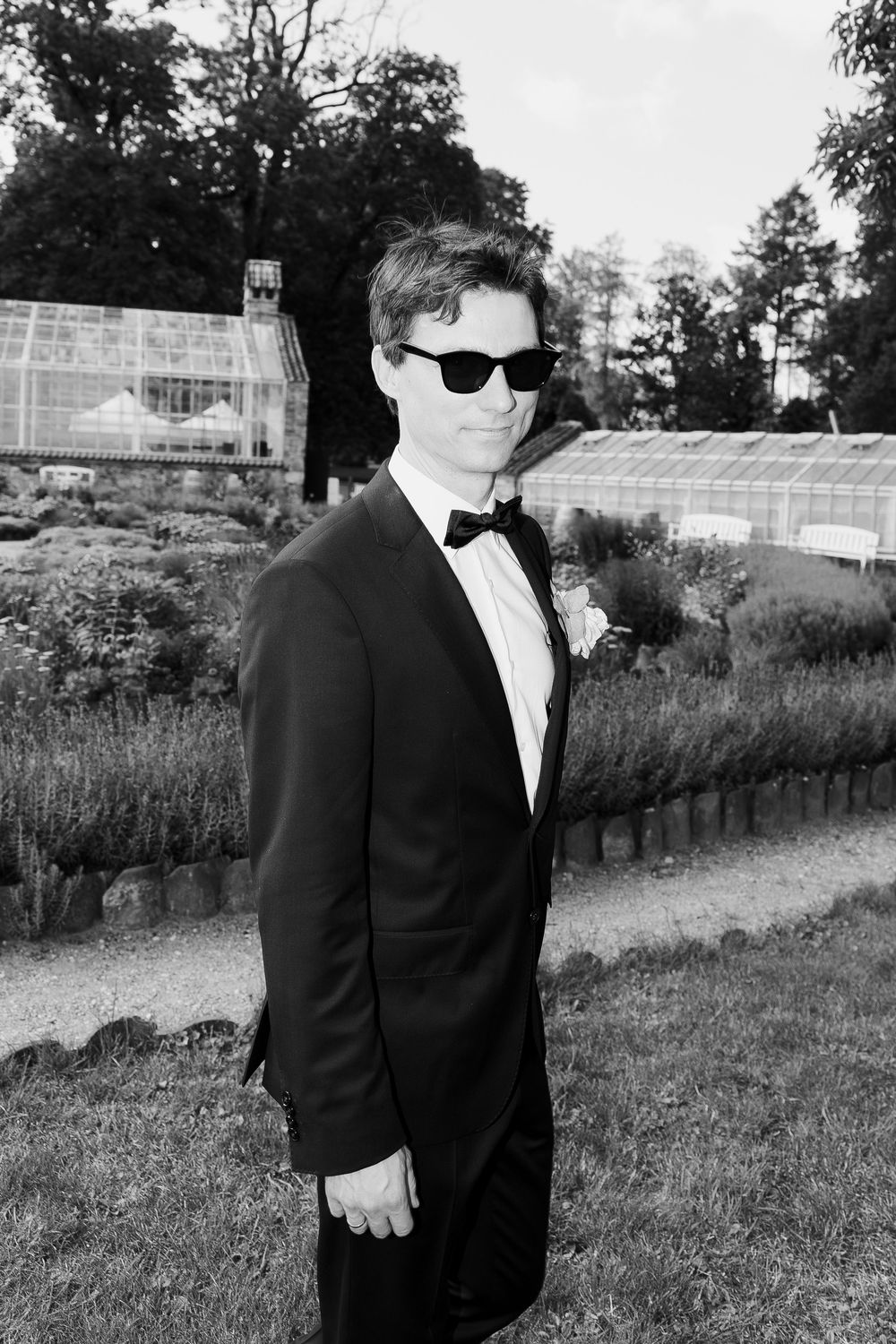 Groom in black tuxedo and sunglasses poses in outdoor garden setting.