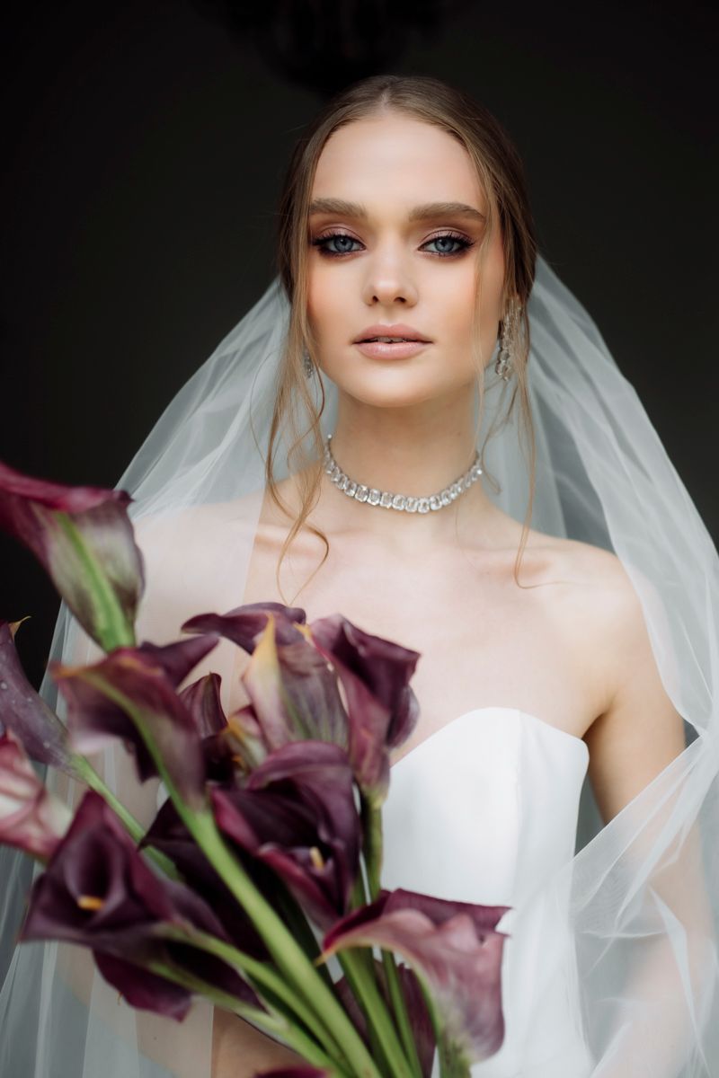 Beautiful bride wearing diamond necklace and veil holds dark purple calla lily bouquet in stunning bridal portrait.
