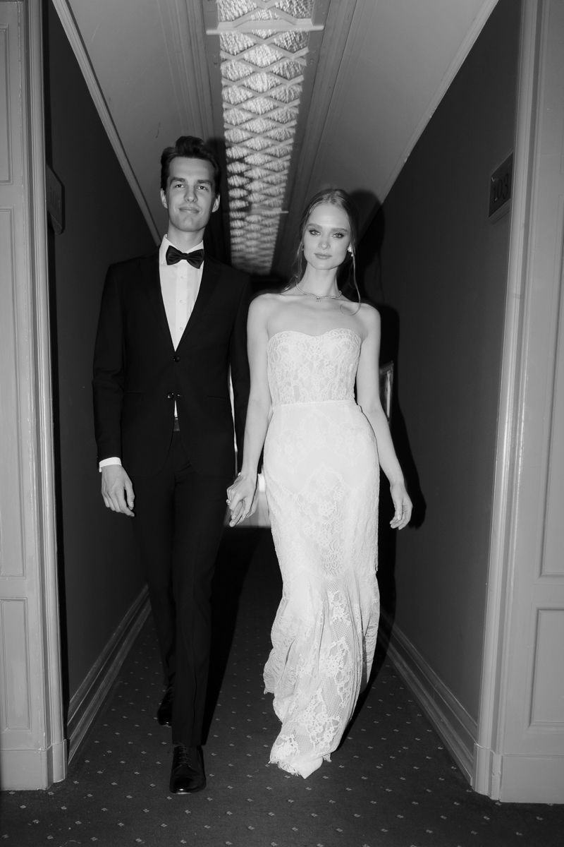 Elegant couple in wedding attire walking down a hallway, groom in tuxedo and bride in lace gown, black and white photo.