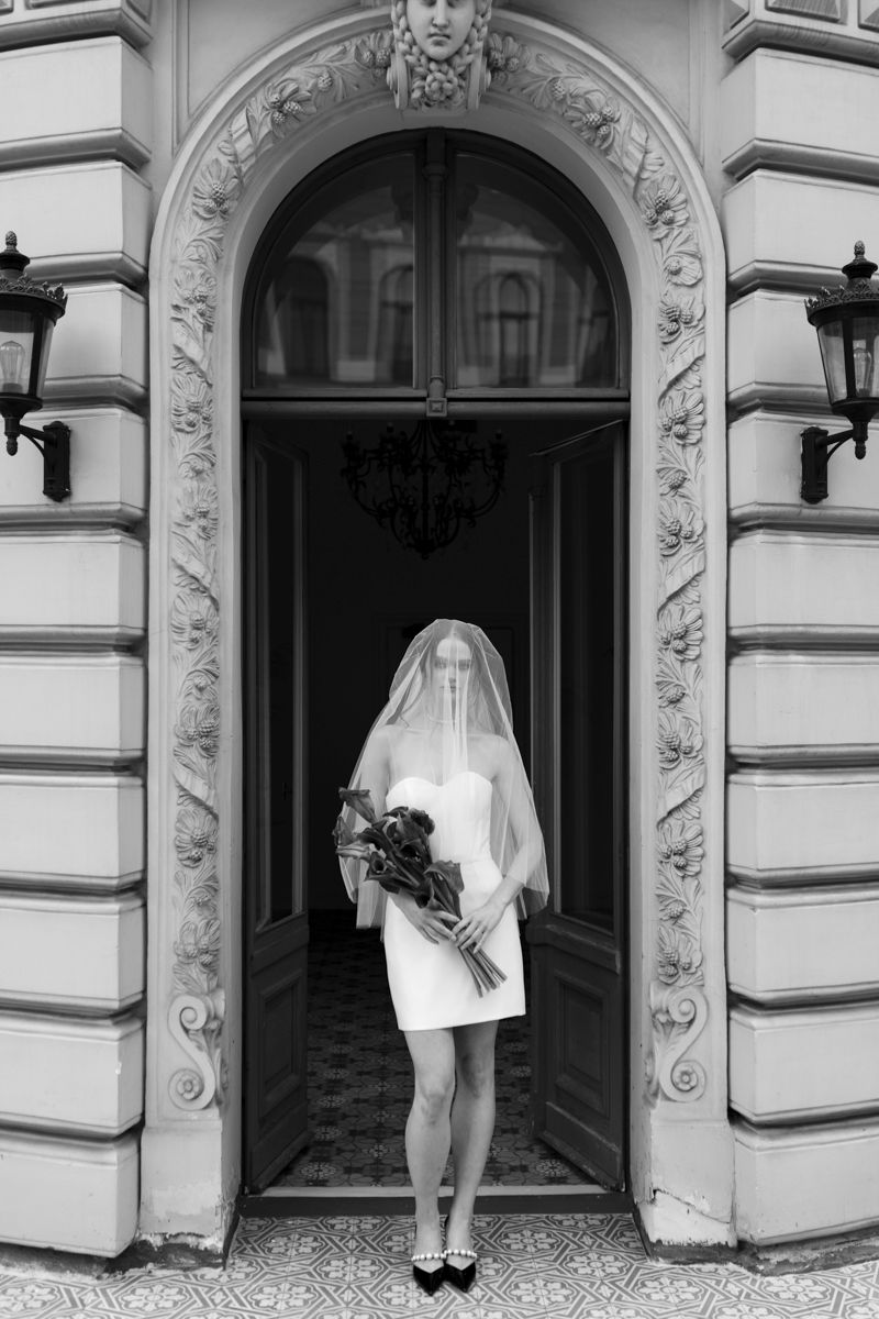 Bride in mini dress with face-covering veil holds bouquet in arched ornate building entrance, B&W.