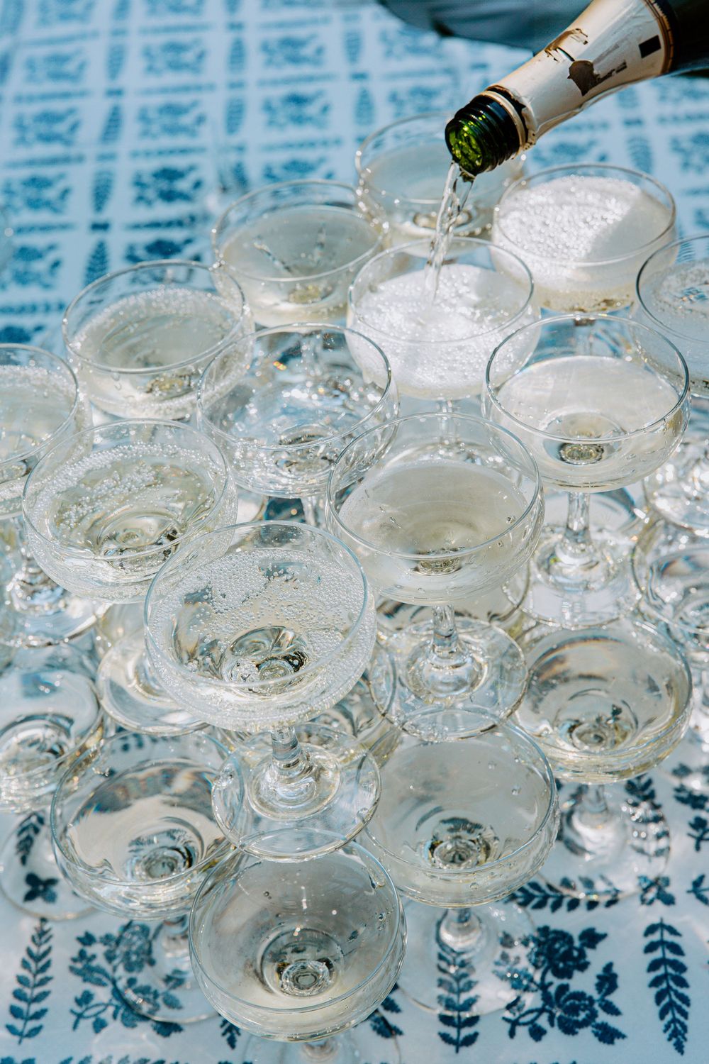 Champagne being poured into elegant coupe glasses arranged on a patterned blue and white tablecloth.
