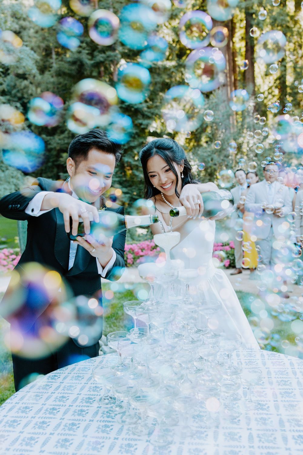 A couple in wedding attire playfully poses with bubbles floating around them in an outdoor garden setting.