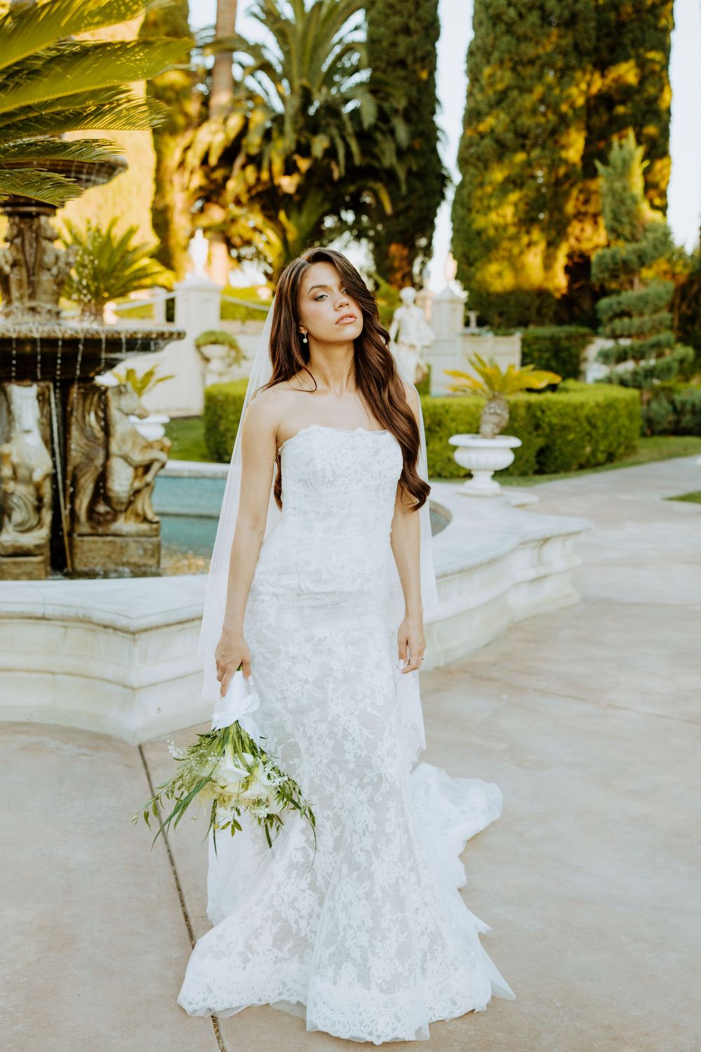 A bride in a flowing white lace wedding dress twirls gracefully in a garden setting with palm trees at sunset.