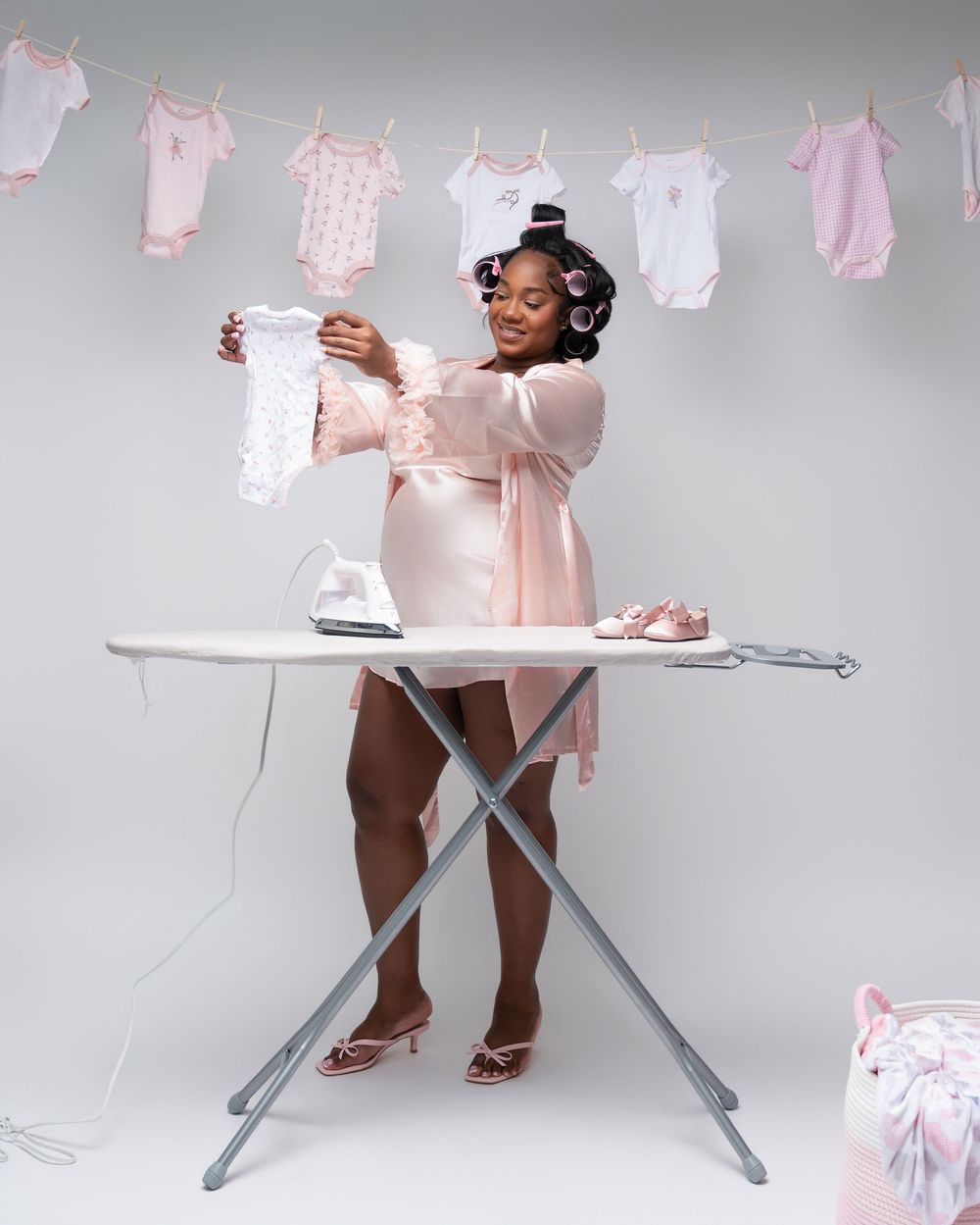 Pregnant woman in pink robe with hair rollers ironing baby clothes near hanging pink onesies garland.
