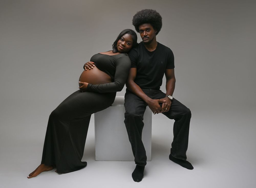 Expecting couple in matching black outfits pose for maternity photo shoot against gray studio background.