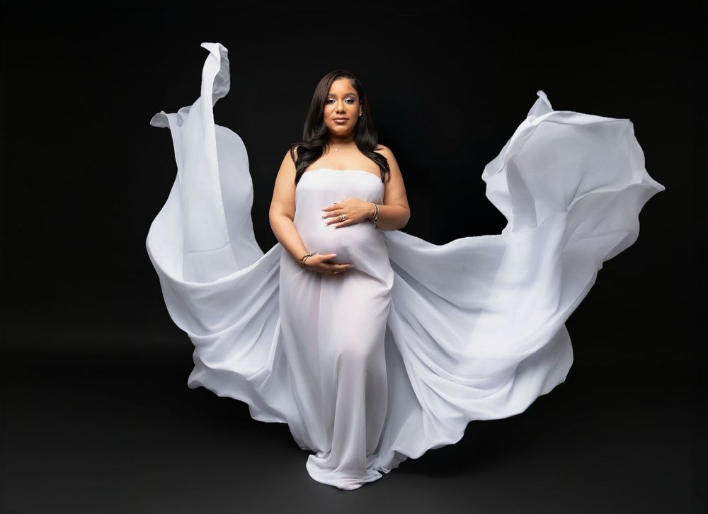 Pregnant woman in flowing white maternity gown poses gracefully against black background with fabric billowing dramatically.