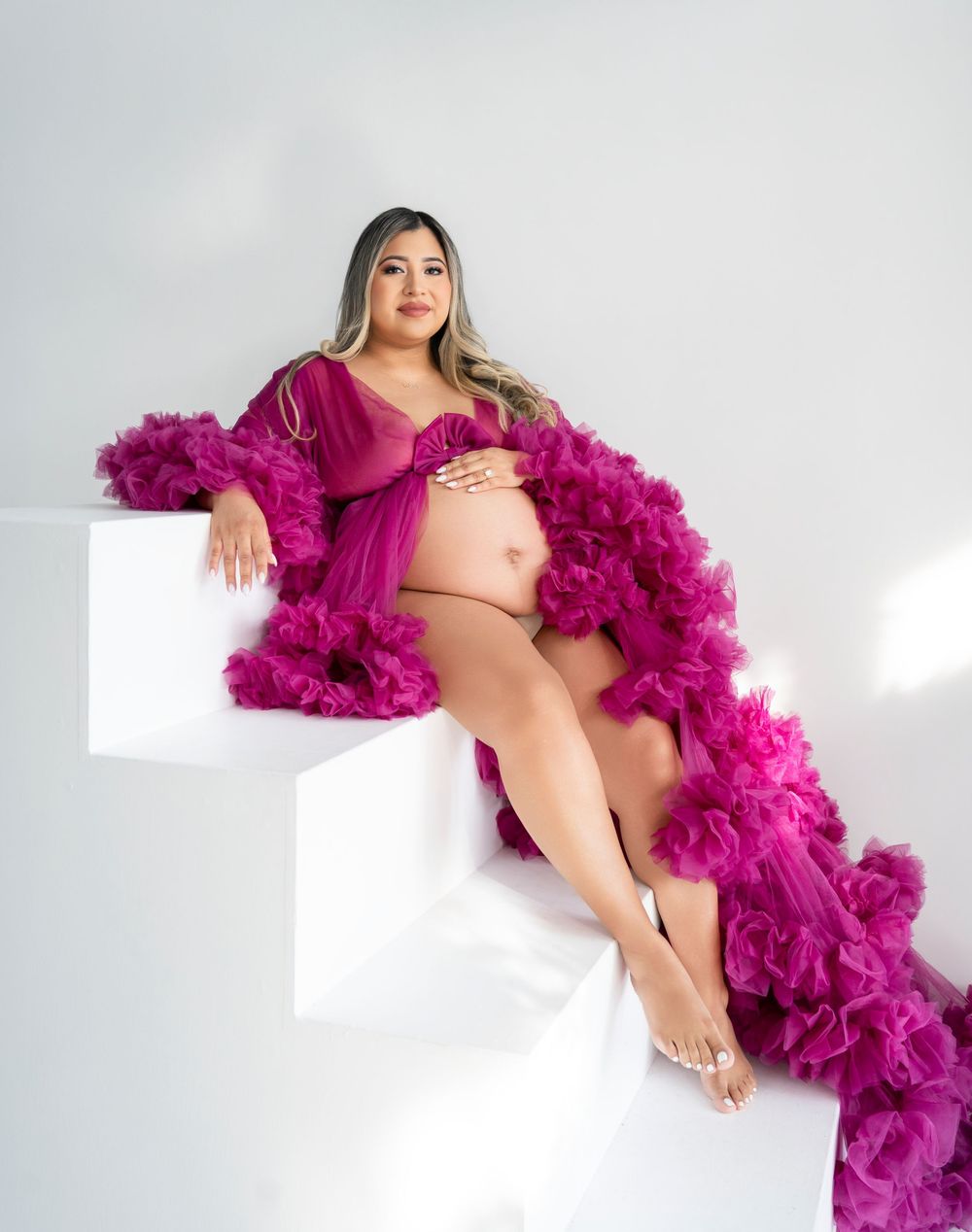 A maternity photo featuring a dramatic magenta tulle robe on white steps in a minimalist setting.
