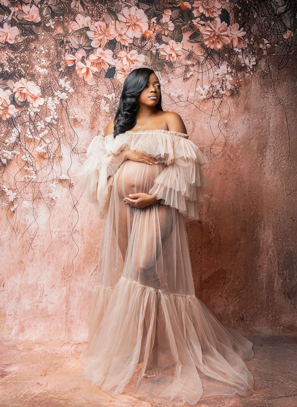Pregnant woman in sheer blush tulle maternity gown posing against floral pink backdrop for professional photo shoot.
