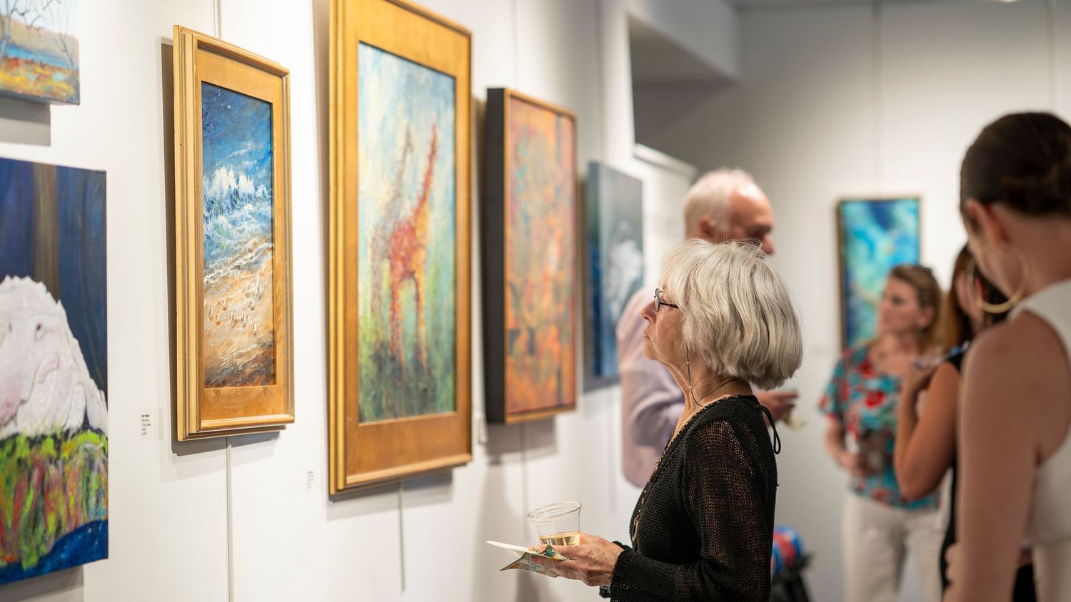 Art gallery visitors view colorful paintings displayed on white walls during an exhibition opening.