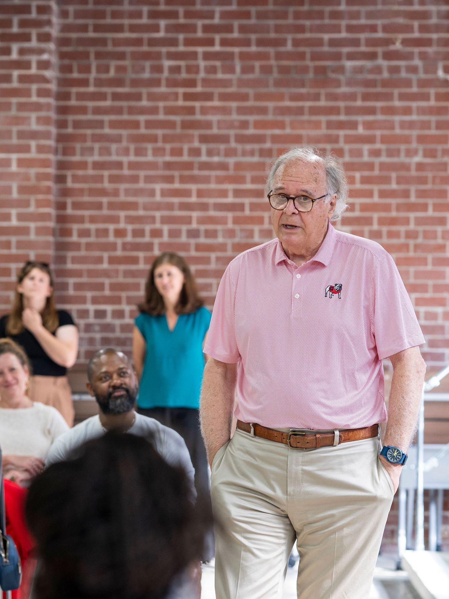 A group gathers in a room with brick walls as someone in a pink shirt addresses them during a meeting.