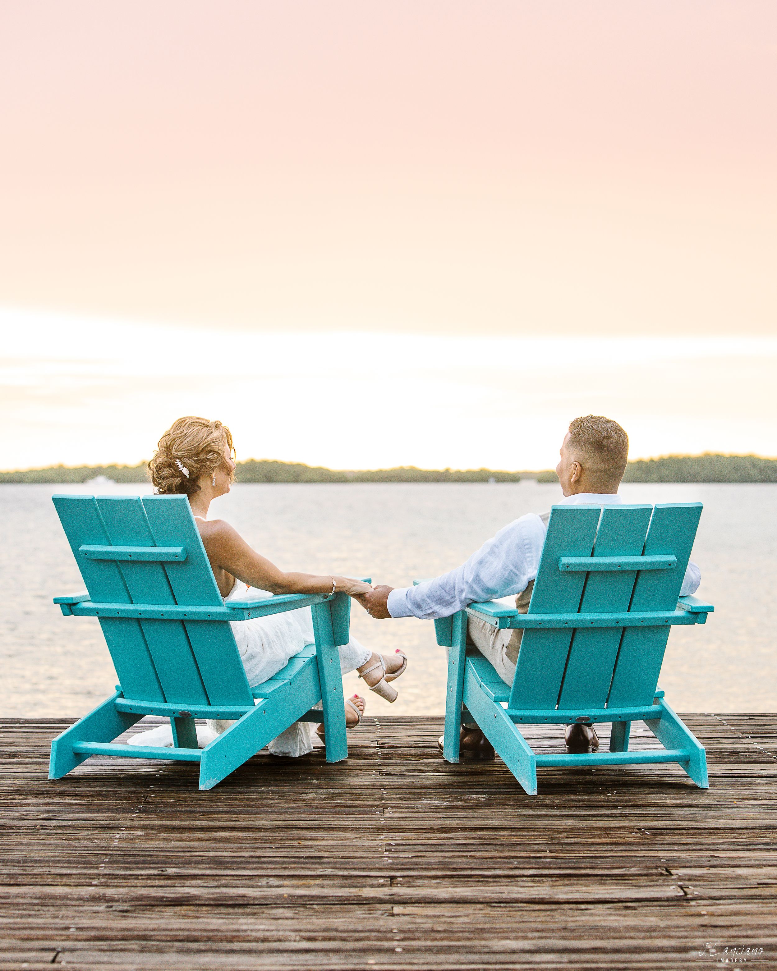 Two turquoise Adirondack chairs on a wooden dock overlooking calm waters at sunset.