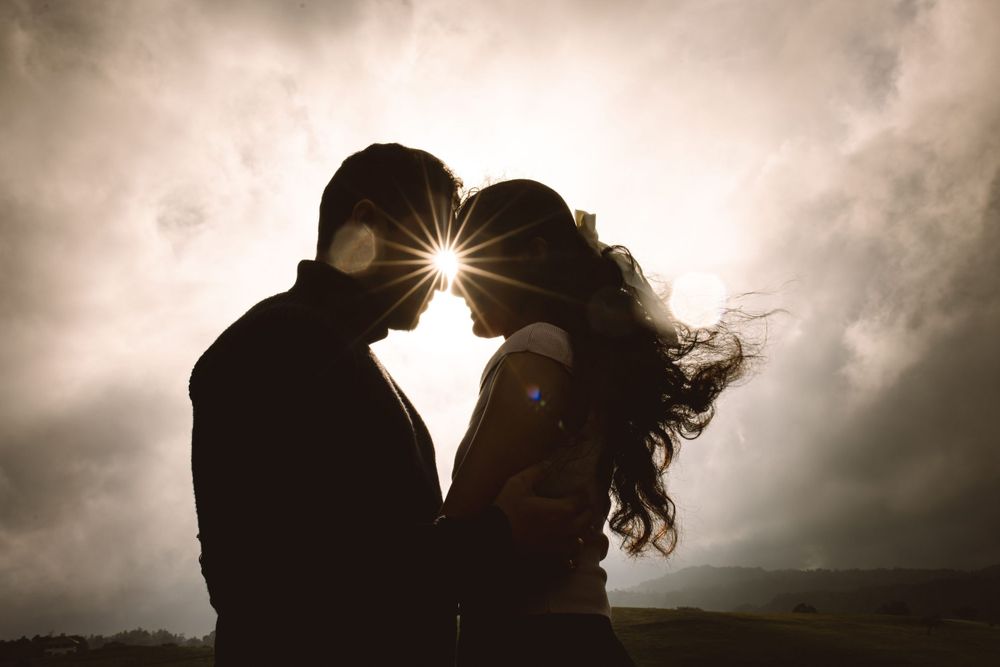 Dramatic silhouettes of a couple standing on a hilltop against a cloudy sky with sun rays breaking through.