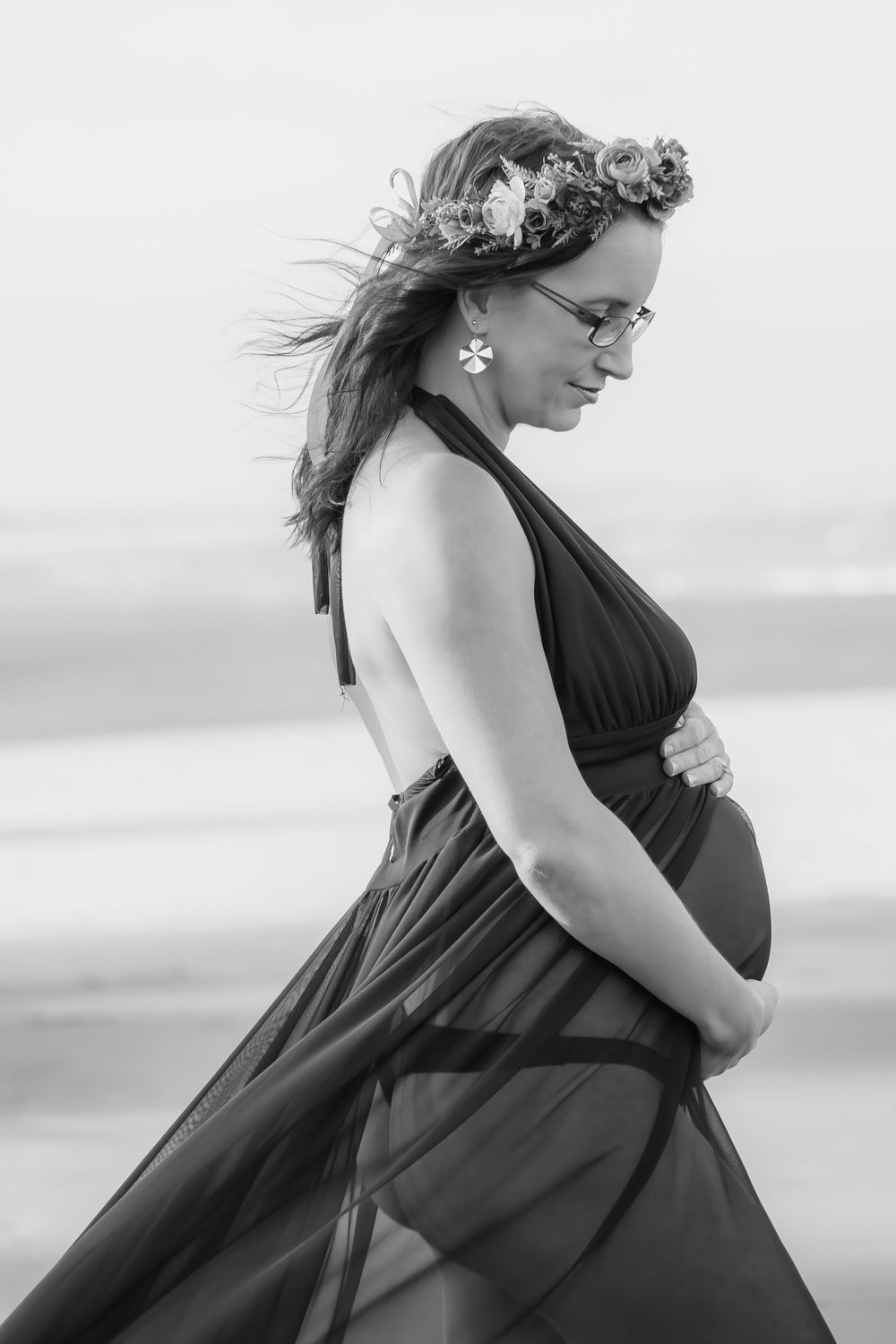 A black and white maternity photo shows an expectant mother in a flowing dress and floral crown on the beach.