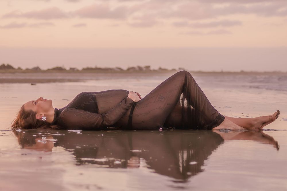 A maternity photo at sunset shows a woman lying on wet sand at the beach.