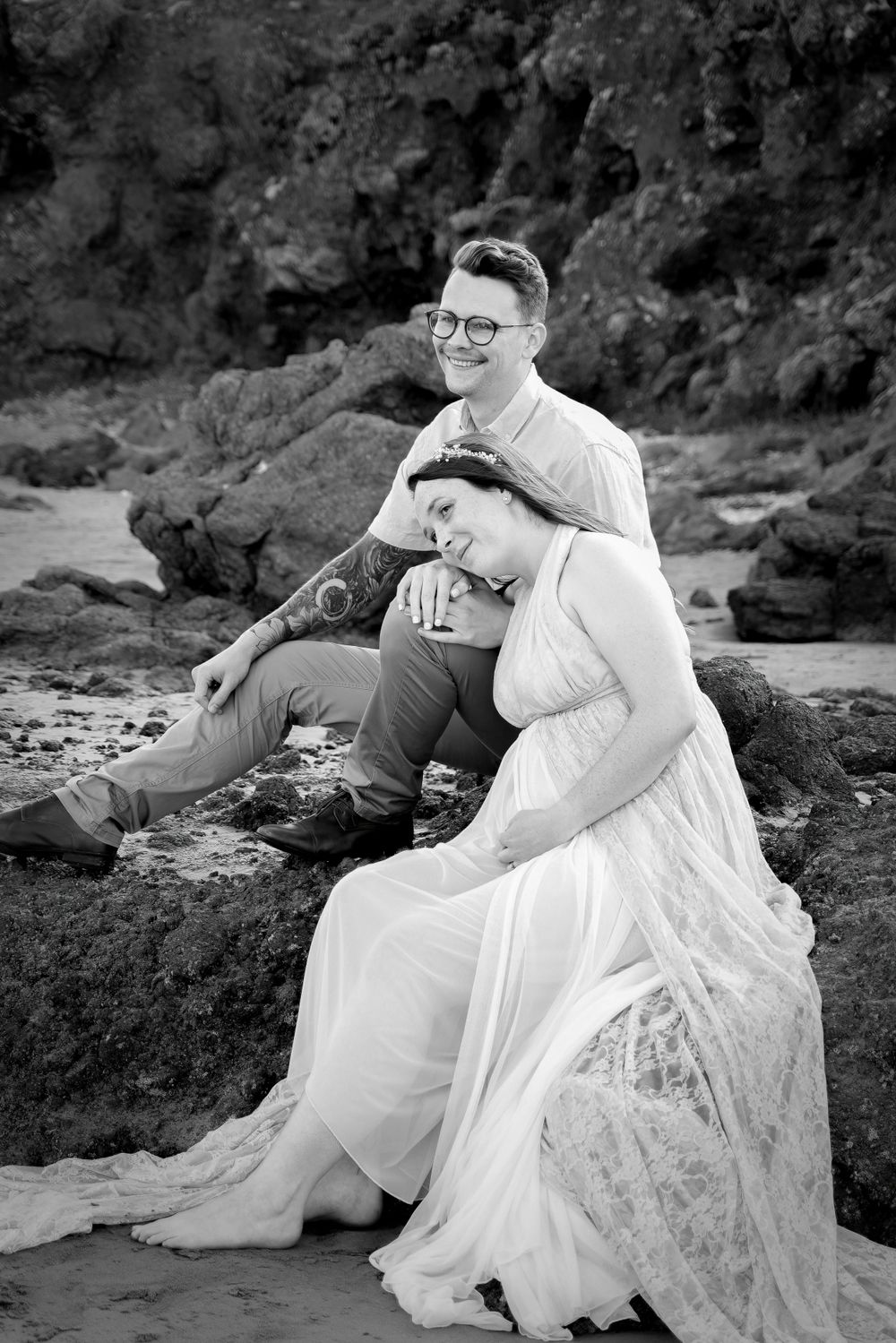 A black and white beach photo shows a couple sitting together on coastal rocks.