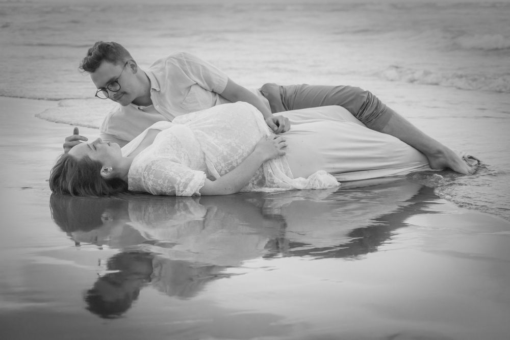 A romantic black and white photo shows a couple lying in shallow beach water.