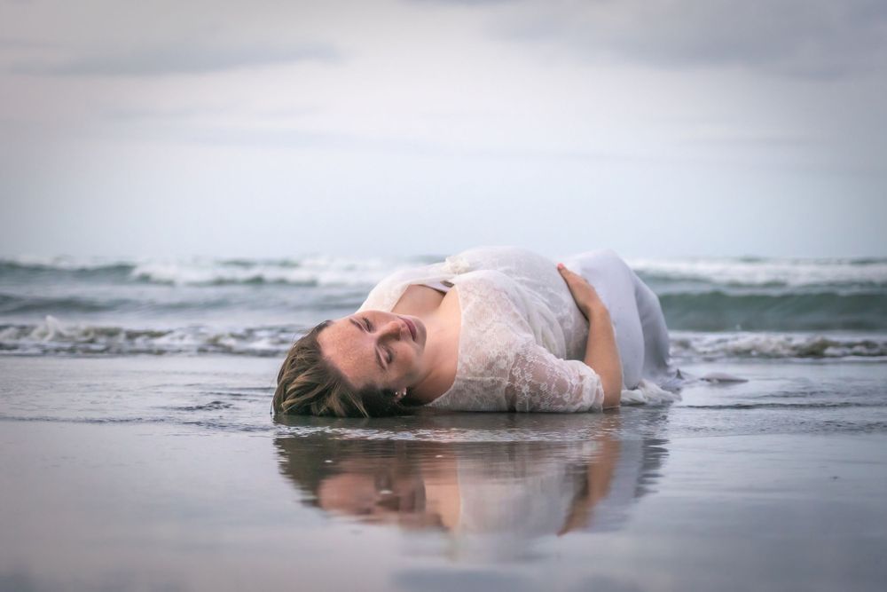 A dreamy maternity photo shows a woman in white lying in shallow ocean waves.