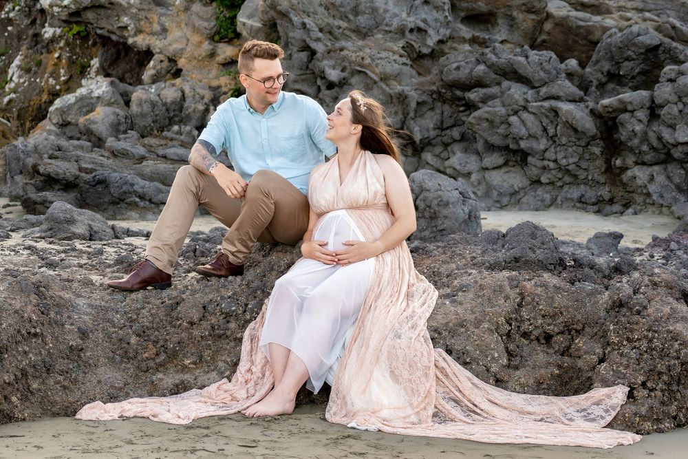 A couple in light colored clothing sits on rocky coastal terrain during a maternity photoshoot.