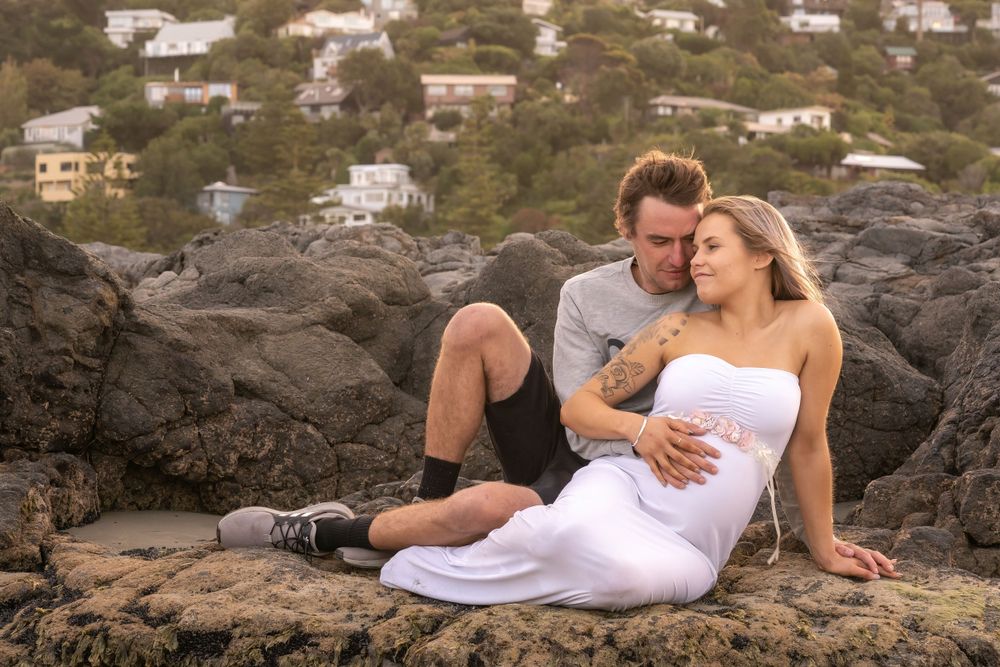 A romantic couple rests on volcanic rocks by the ocean with coastal homes visible in the background.