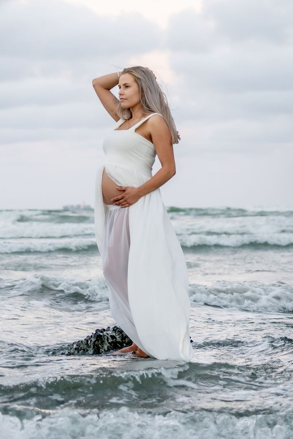 A maternity photoshoot featuring flowing white fabric against a dramatic ocean backdrop.