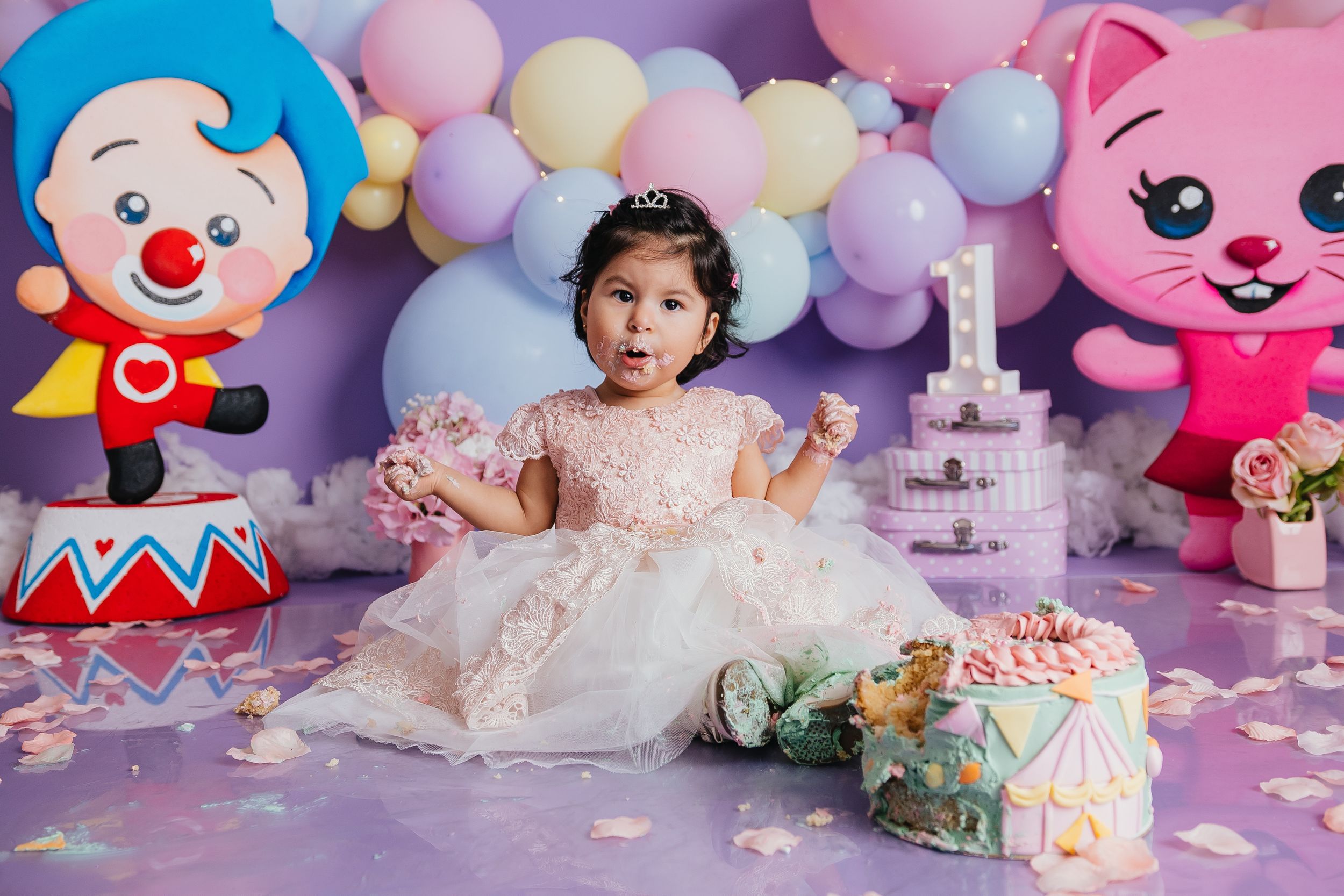 A festive first birthday celebration setup with colorful balloons, plush toys, and a pink theme against a purple backdrop.