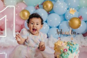 Colorful first birthday celebration setup with balloons and cake against pink and blue backdrop.
