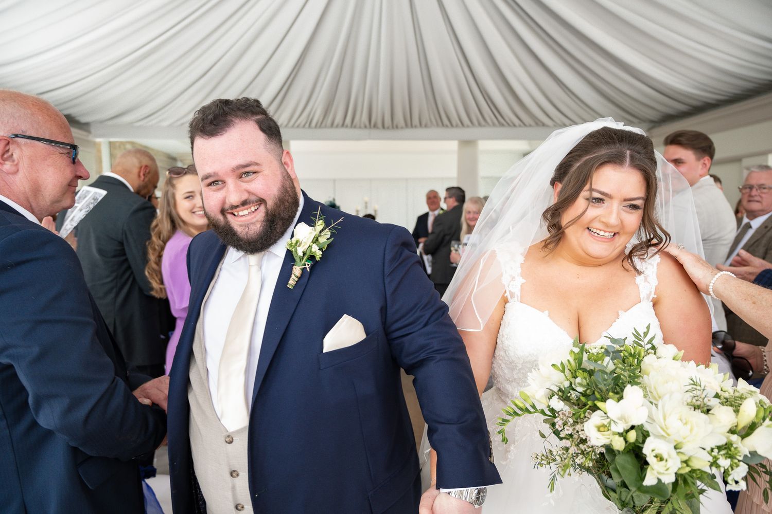 A wedding celebration under a white tent with guests mingling and a bride holding a bouquet of white flowers.