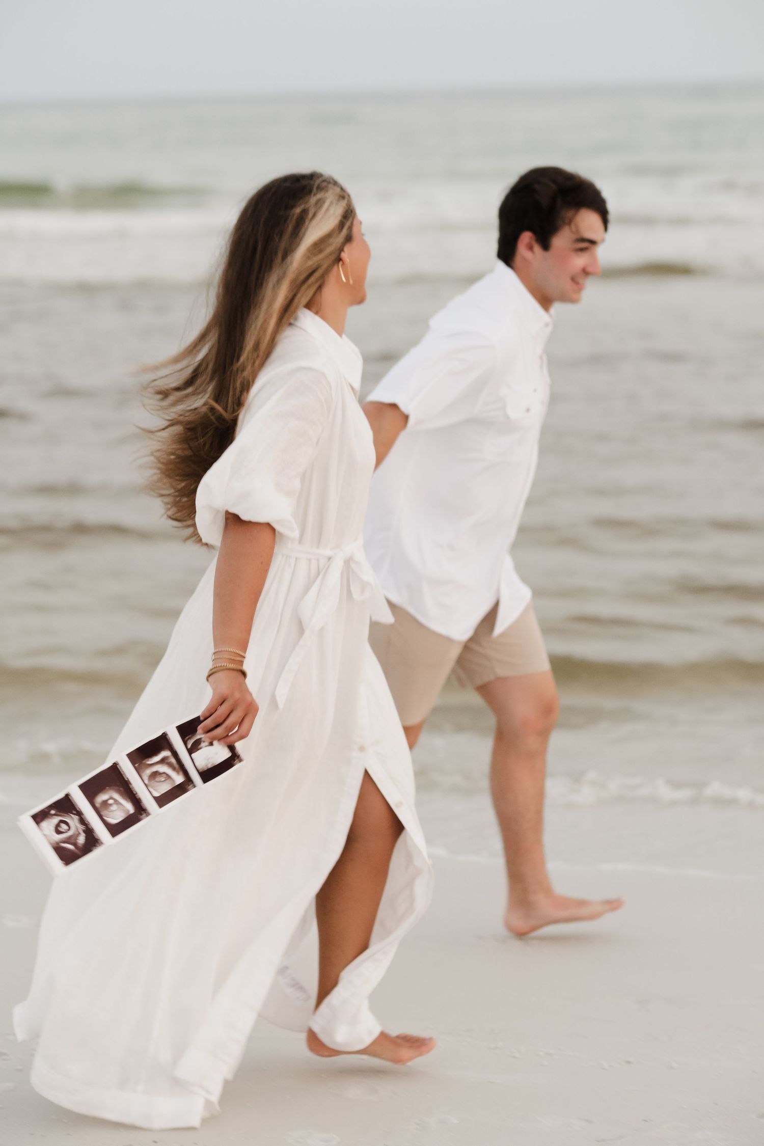 Couple walking on beach holding ultrasound photos in white outfits.