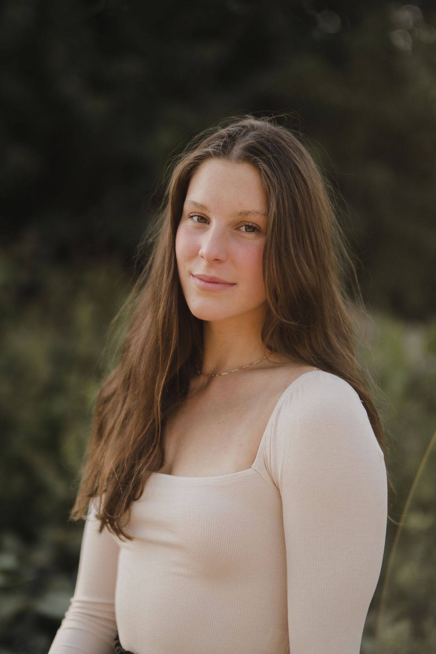 Person in cream colored long sleeve top poses against natural outdoor backdrop.