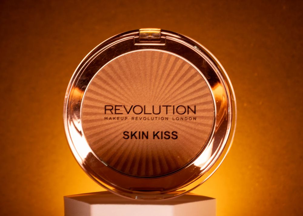 Makeup compact with 'Revolution Skin Kiss' text photographed with warm amber lighting.