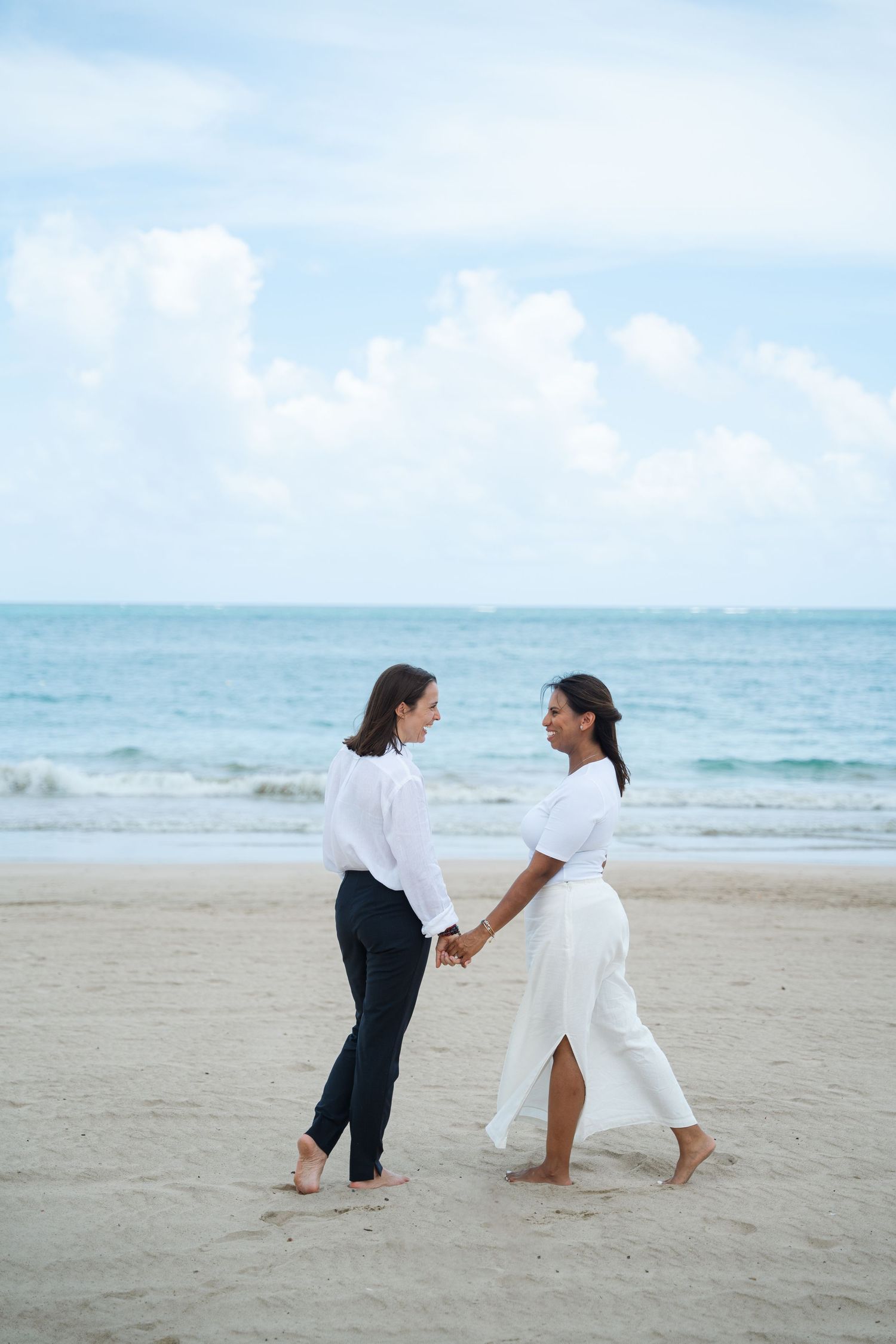 Two people in white outfits hold hands while walking barefoot on a serene beach with turquoise waters and blue sky.