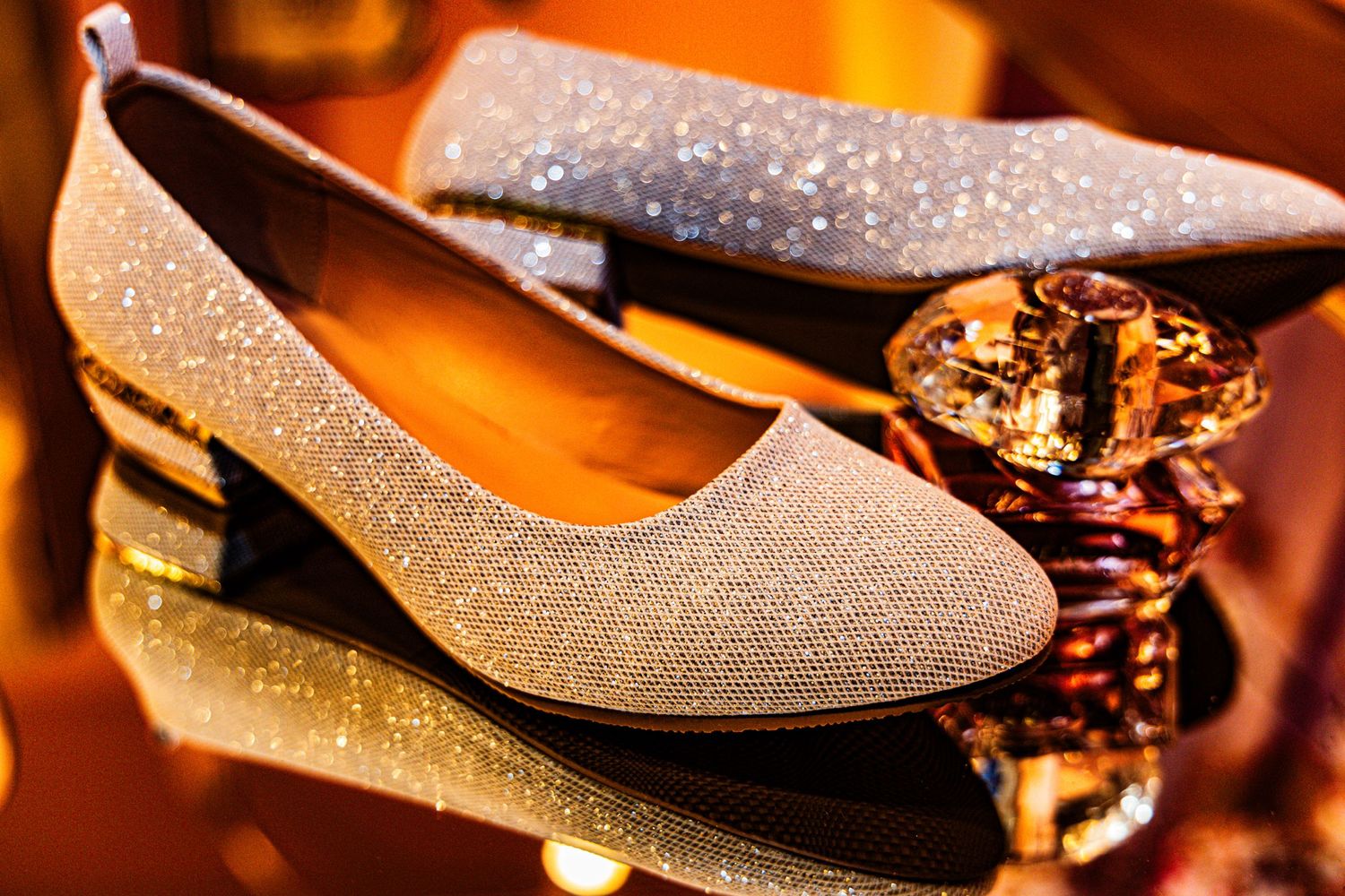 Sparkling silver glitter low-heel bridal flats reflected on glossy surface with golden bokeh background.