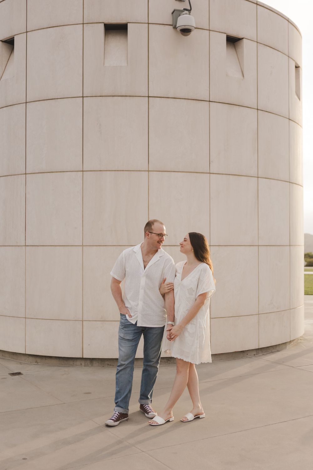 A couple in coordinating white outfits stands beside a curved concrete wall outdoors.