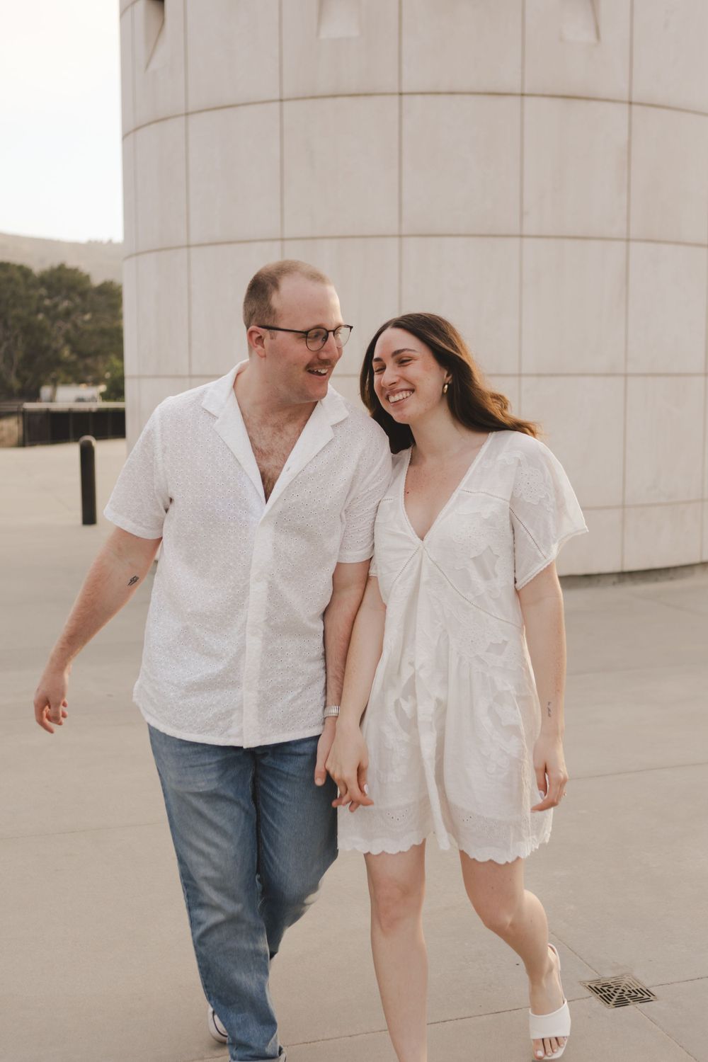 A couple shares a joyful moment while walking near a curved white building exterior.