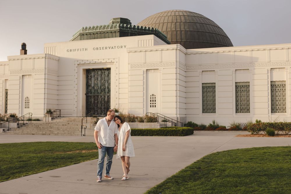 A couple strolls in front of the Griffith Observatory building at sunset.