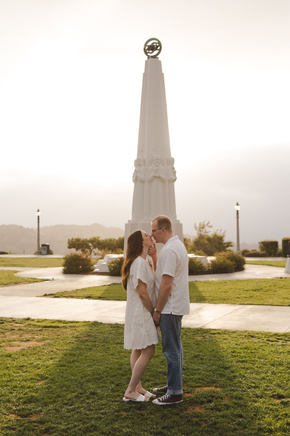 A couple shares a romantic moment in front of a tall monument on a grassy lawn.