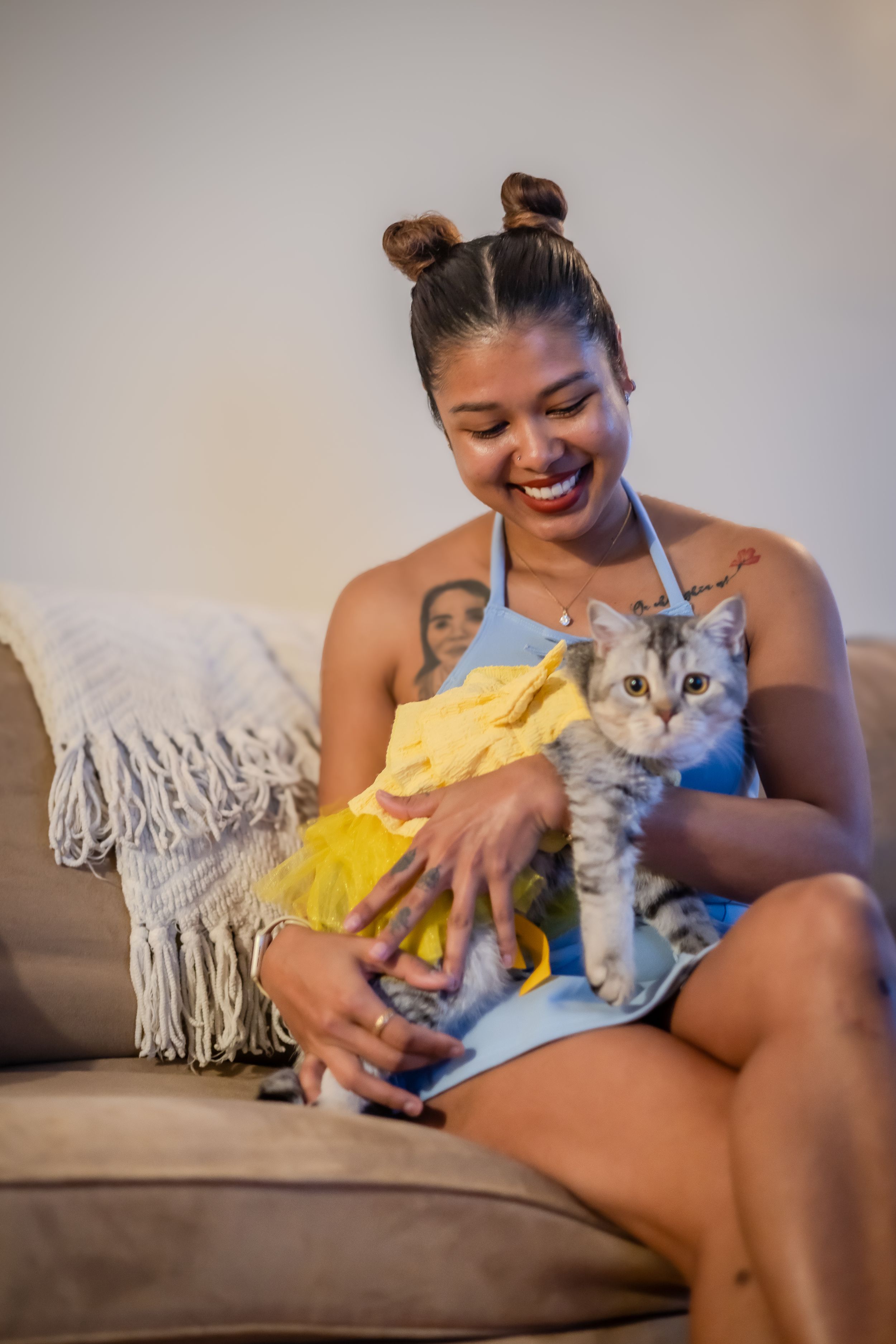 A joyful moment on the couch with a gray cat wearing a yellow sweater as its owner laughs warmly.