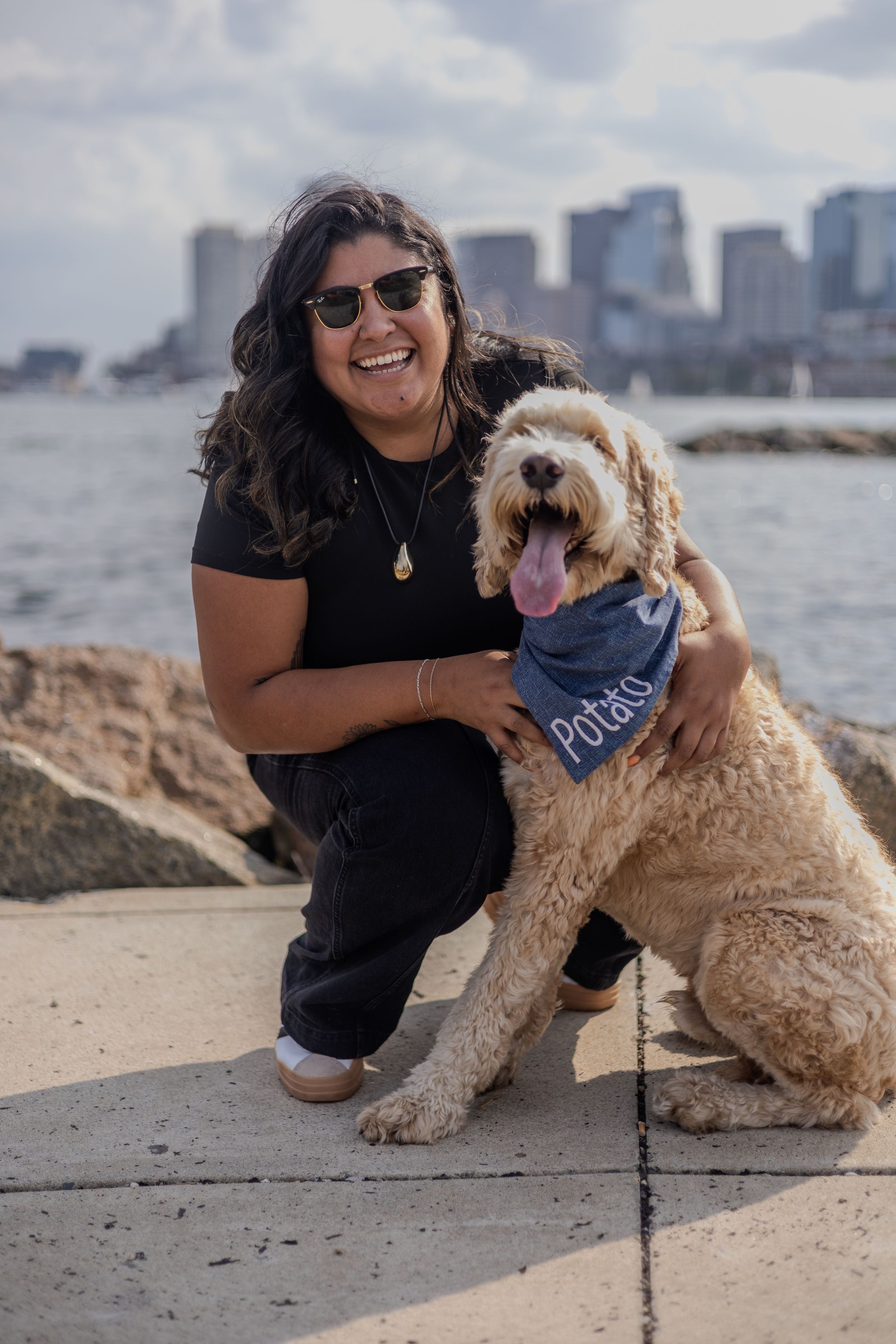 A happy person and their goldendoodle dog posing together by the Boston waterfront.
