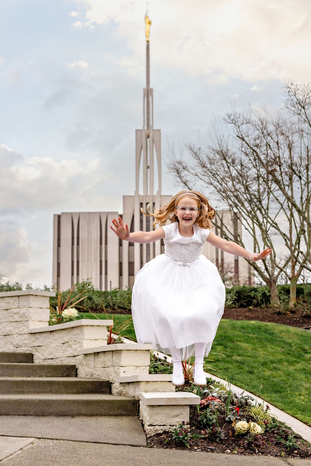 A joyful figure in a white dress leaps down stone steps in front of a Mormon temple with a tall spire.
