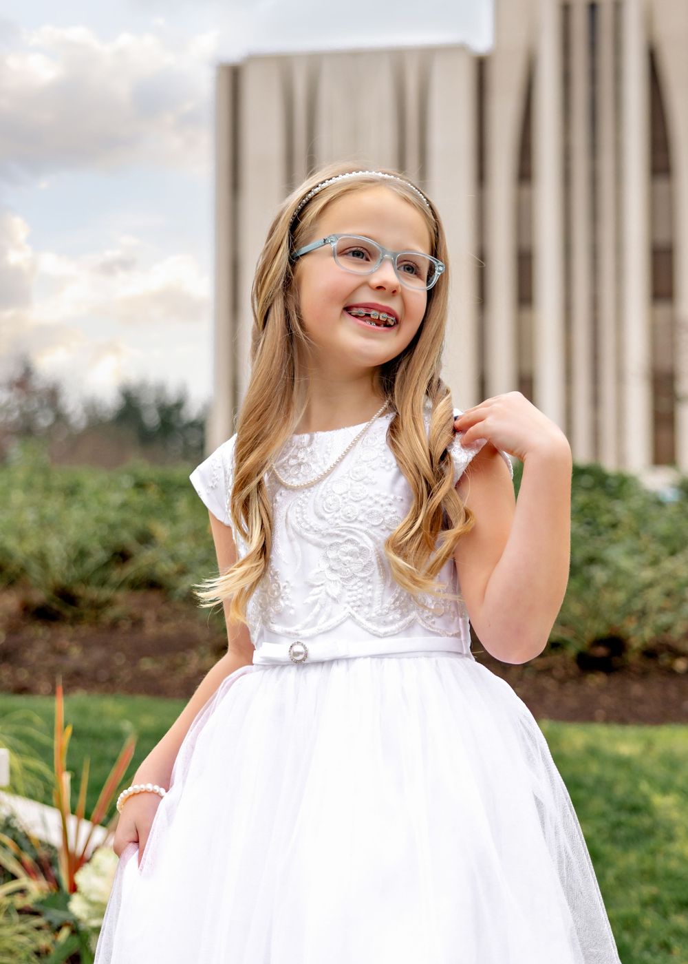A young child in a white lace dress poses in front of a building on a grassy area.