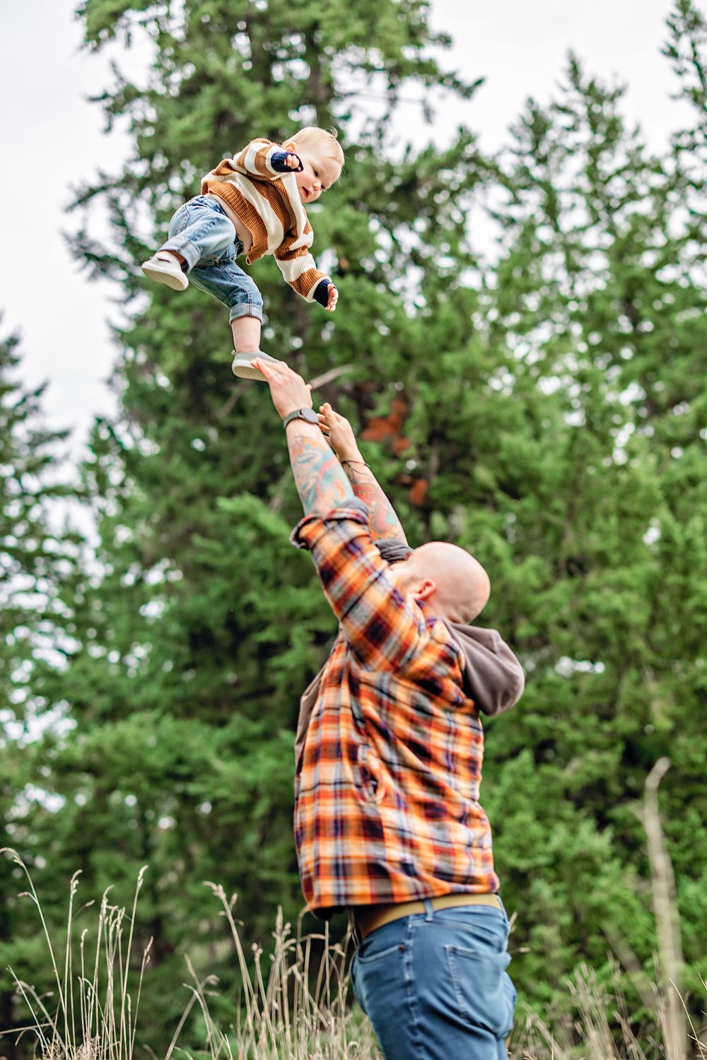 A toddler in plaid shirt and jeans plays joyfully outdoors against a backdrop of evergreen trees.