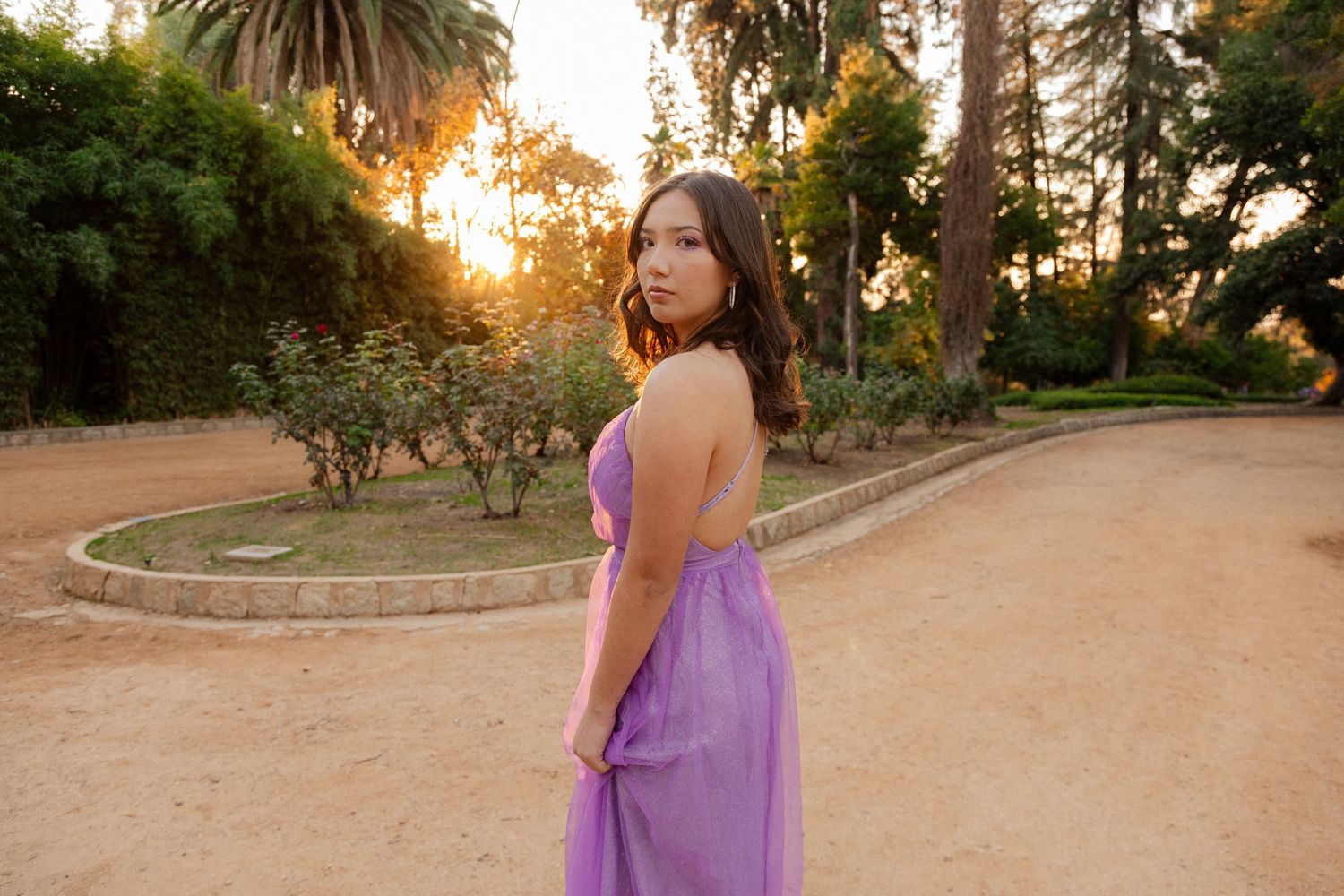 A woman in purple dress poses in a garden path at golden hour.