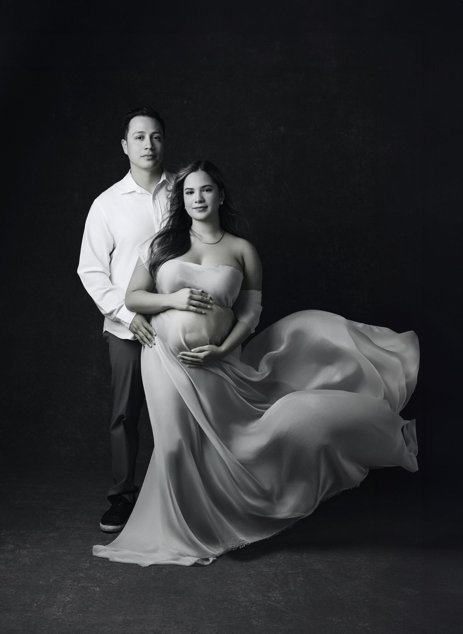 Elegant maternity photo with flowing dress against dark backdrop.