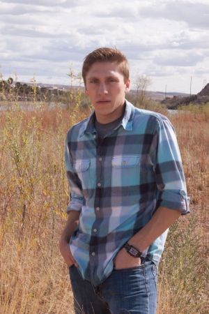 Person in blue plaid shirt and jeans poses in outdoor field with tall grass and cloudy sky in background.