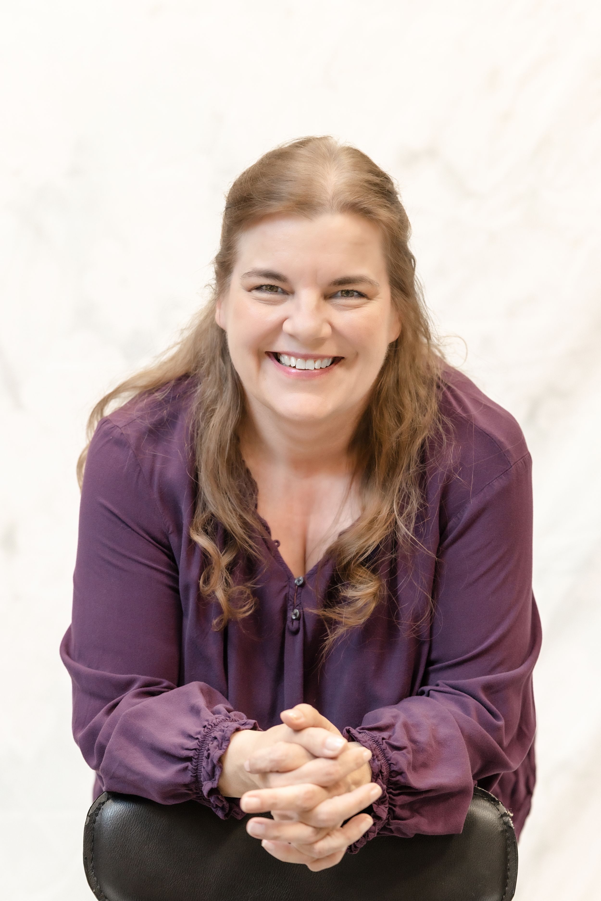 A person in a purple top leaning on chair back while smiling warmly at the camera in a photo studio setting.