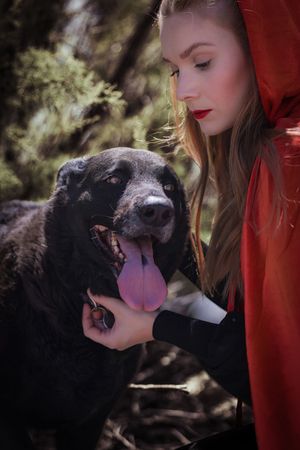 A black dog with its tongue out being petted while sitting next to someone wearing a red hood in a forest setting.