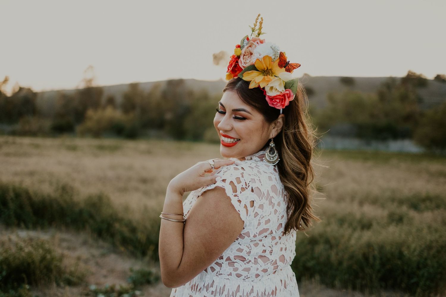 Woman in a white lace dress with a colorful flower crown and dangling earrings, smiling outdoors in a field.