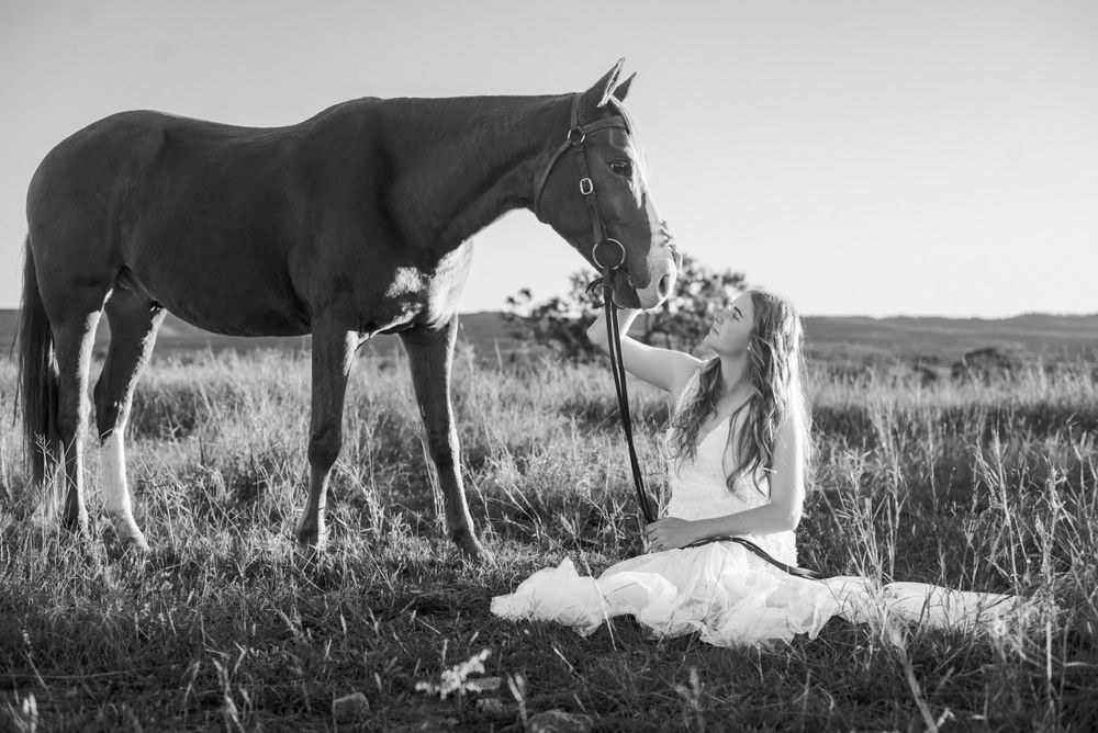 A black and white photo of someone in a wedding dress lying on grass near a standing horse.