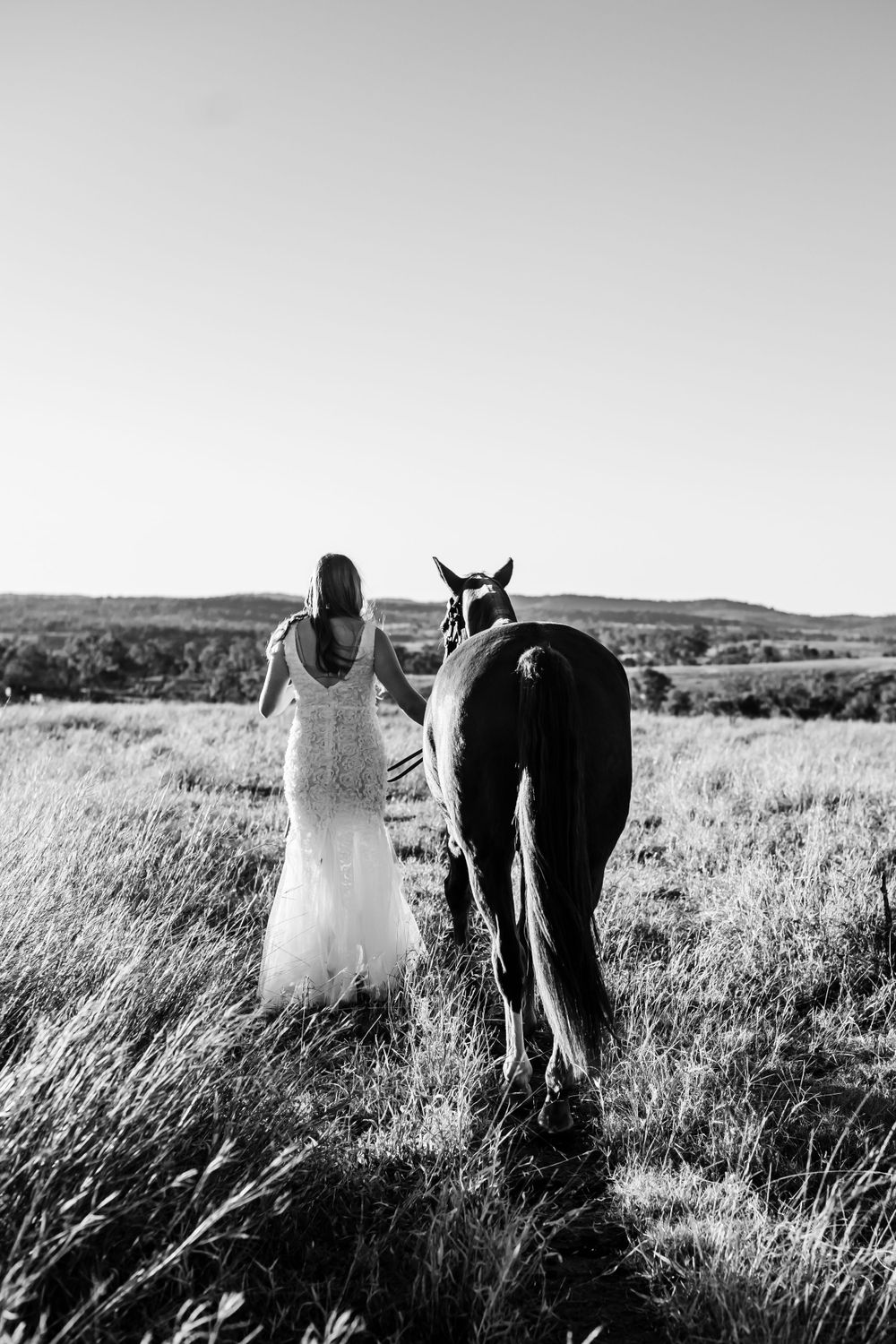 Person in white dress walking alongside a dark horse through a grassy meadow in black and white.